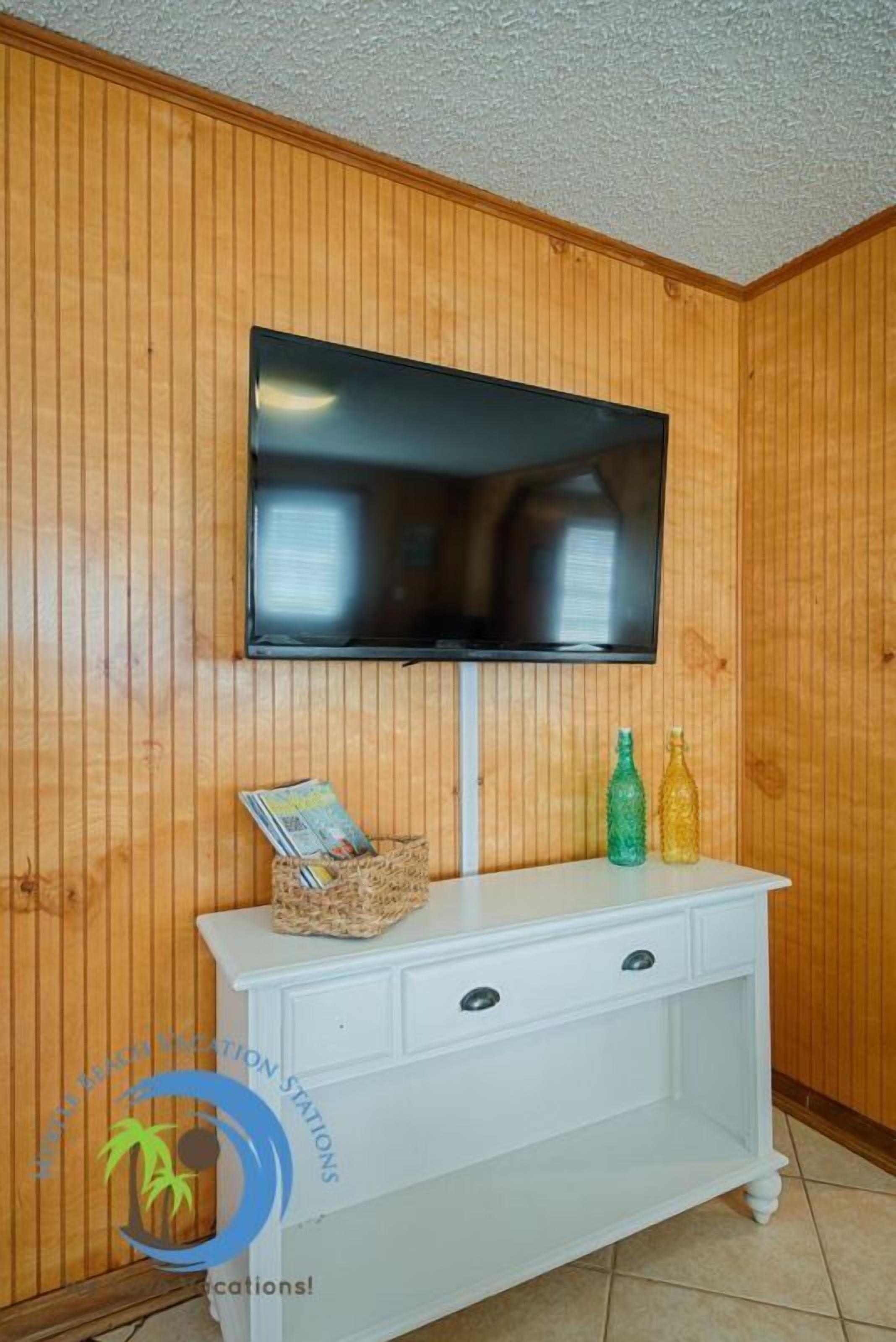 A room with wooden panel walls, a mounted flat-screen TV, a white console table with magazines and bottles, and a logo for "MALIBU RESORTS & RECREATION STATIONS".
