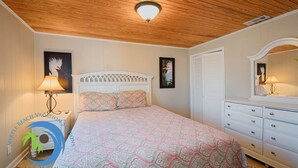 Quarto com teto de madeira, cama com roupa de cama estampada, mesa de cabeceira com abajur, cômoda e espelho.