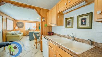 A compact kitchen with wooden cabinets, a granite countertop, and a sink. There is a dining area with a table and chairs, and a living room with a sofa and a decorative wall hanging.