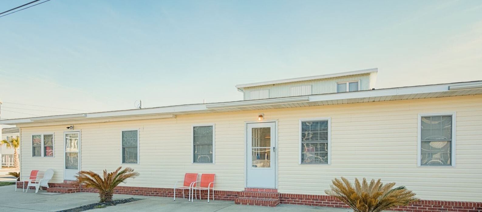 A single-story motel with a flat roof, a front door, and a small porch with two chairs.