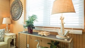 Um quarto aconchegante com painéis de madeira, uma grande janela com persianas, um adorno na parede, uma luminária, uma planta, uma pequena mesa com uma âncora decorativa e uma cesta de vime.