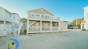 Een vakantiehuis aan het strand met twee verdiepingen, een balkon, een omheind oprit en een heldere lucht.