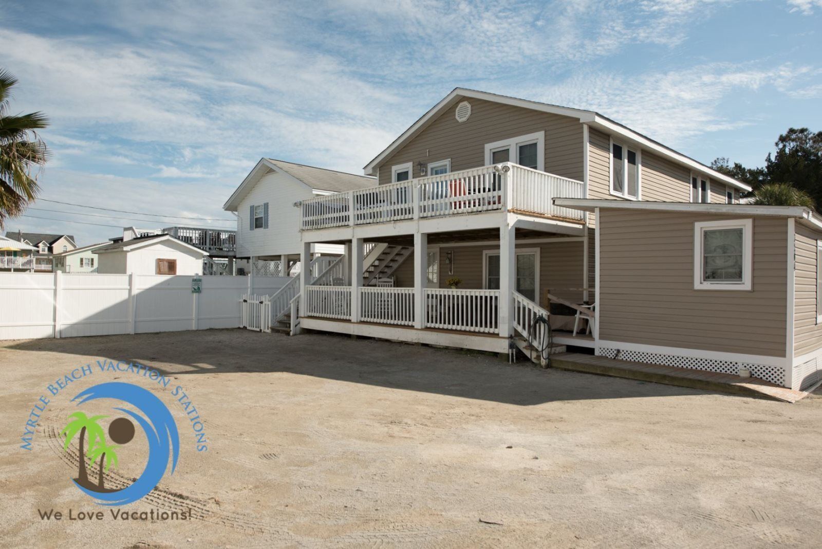 A two-story beach vacation rental with a balcony, a covered porch, and a sandy driveway.