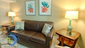 Un canapé en cuir marron agrémenté de coussins à motifs, deux œuvres d’art sur le thème du corail encadrées au mur, et une table d’appoint avec un plateau en verre et une lampe.