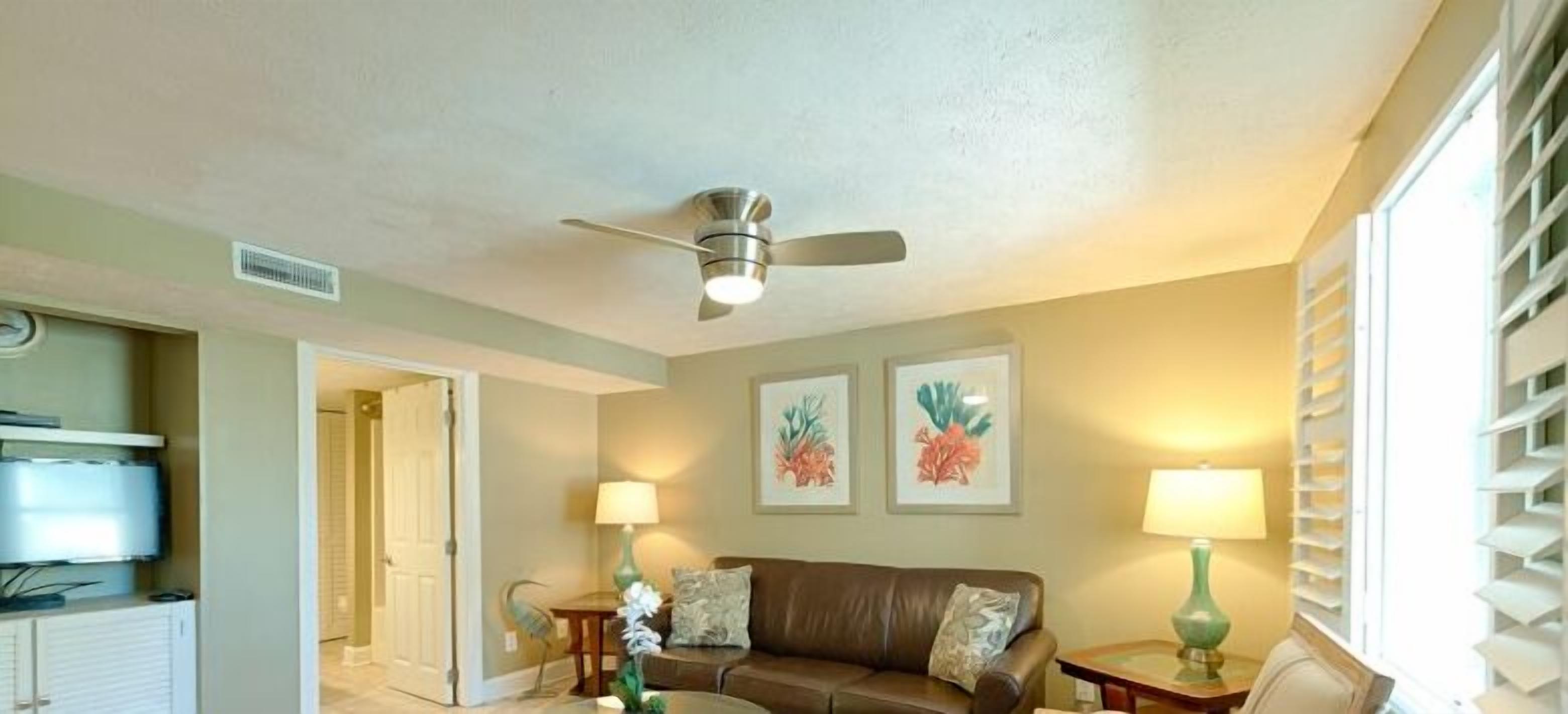 A living room with a ceiling fan, a brown leather sofa, two lamps, and framed floral artwork on the wall.