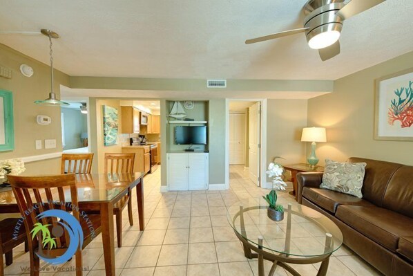 A spacious living area with a dining table, a glass coffee table, a brown leather sofa, and a ceiling fan.