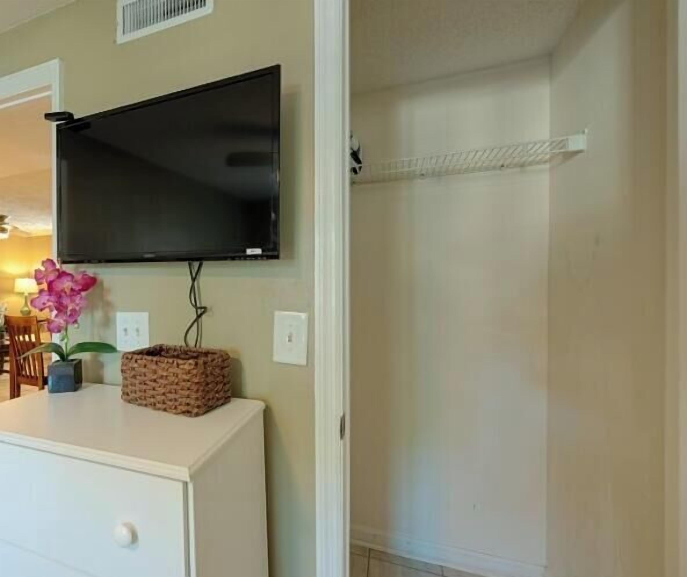 A room with a flat-screen TV mounted on the wall, a small table with a basket, and a white dresser.