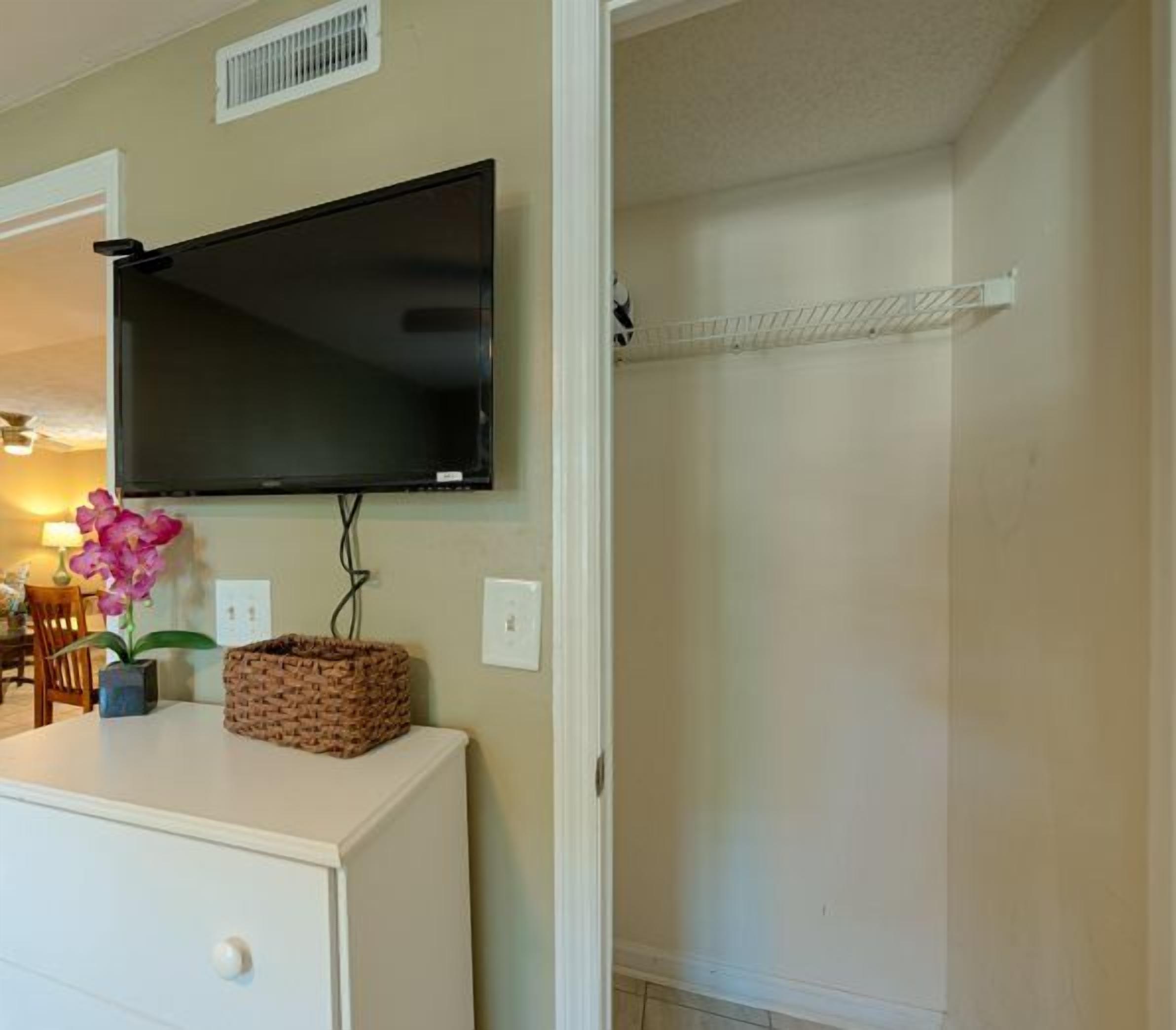 A room with a mounted flat-screen TV, a white cabinet, a wicker basket, and a small potted orchid.