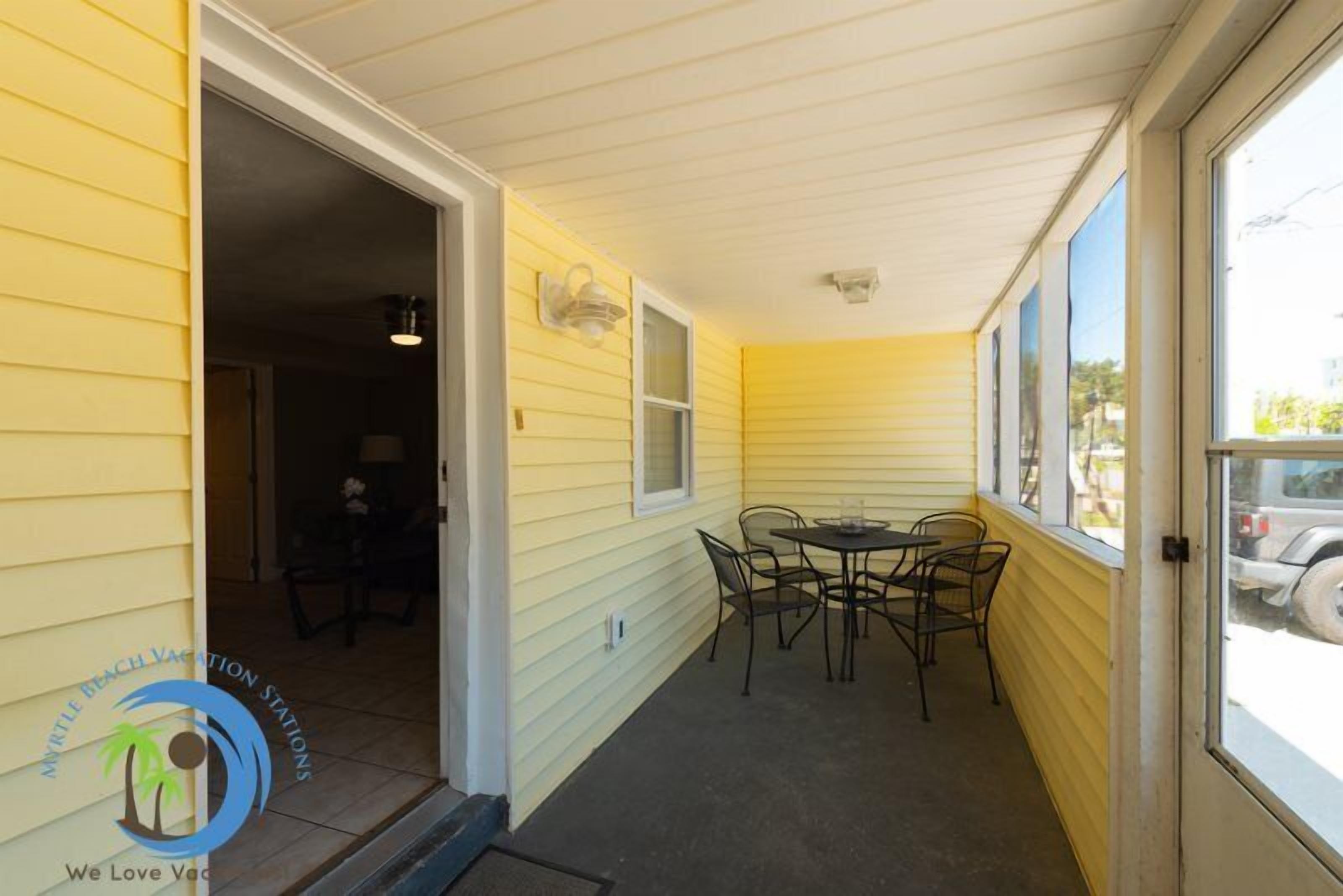 A covered porch with a table and chairs, a door leading to an interior room, and a window with a view of the street.