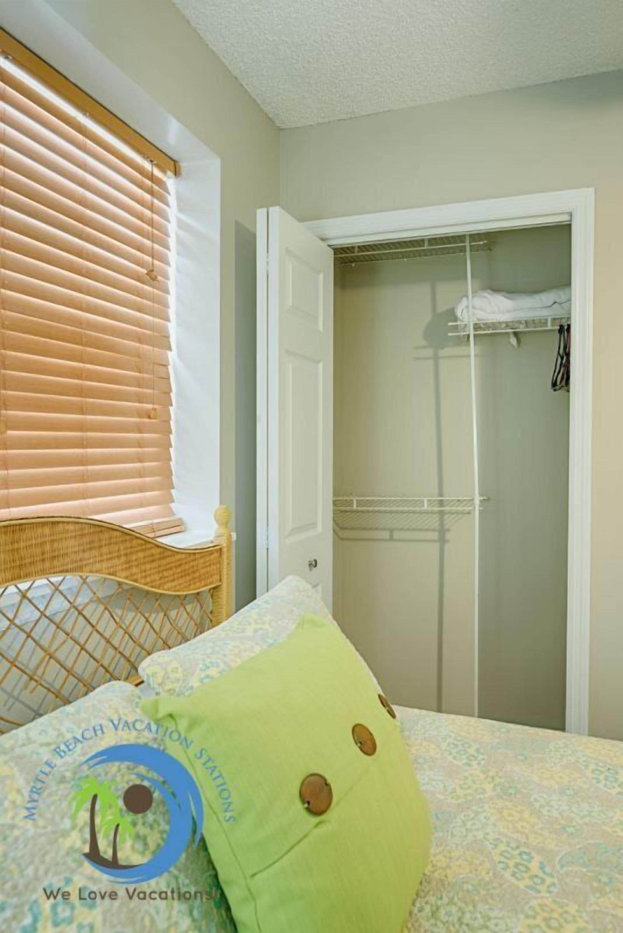 A bedroom with a bed, a window with blinds, a closet with hanging clothes, and a logo on the bedspread.