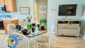A modern kitchen with wooden cabinets, a microwave, and a stove. There is a dining area with a glass table and chairs, a vase with plants, and a television on the wall. - Windy Hill Villa 2 - Beach Retreat (North Myrtle Beach)