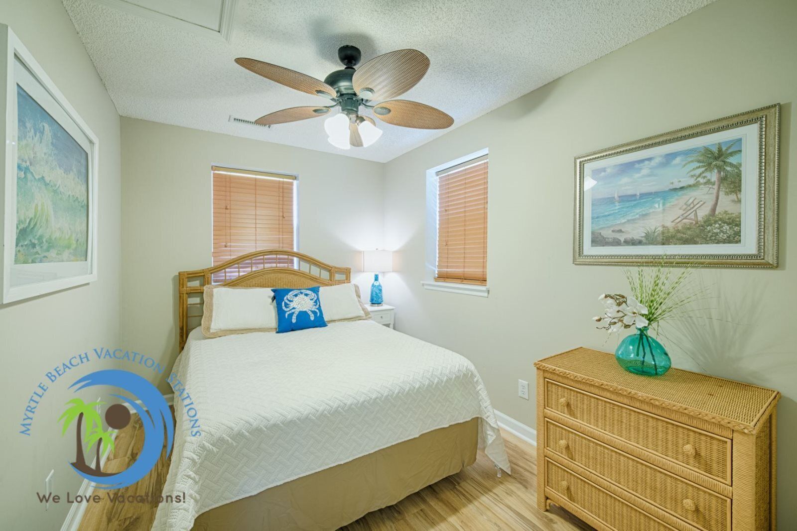 Une chambre à coucher comprenant un lit, un ventilateur de plafond, deux fenêtres avec stores, une table de chevet avec une lampe, une commode en osier et un tableau représentant une plage, encadré.