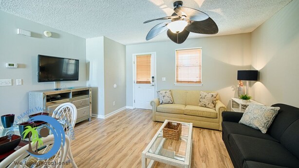 A modern living room with a flat-screen TV, a ceiling fan, a sofa, a coffee table, and a dining area with a white chair and a blue table. - Windy Hill Villa 2 - Beach Retreat (North Myrtle Beach)