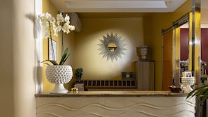 A reception area with a marble counter, a sunburst wall decoration, and a white orchid vase. - Hotel Riva Del Sole (San Vito Lo Capo)