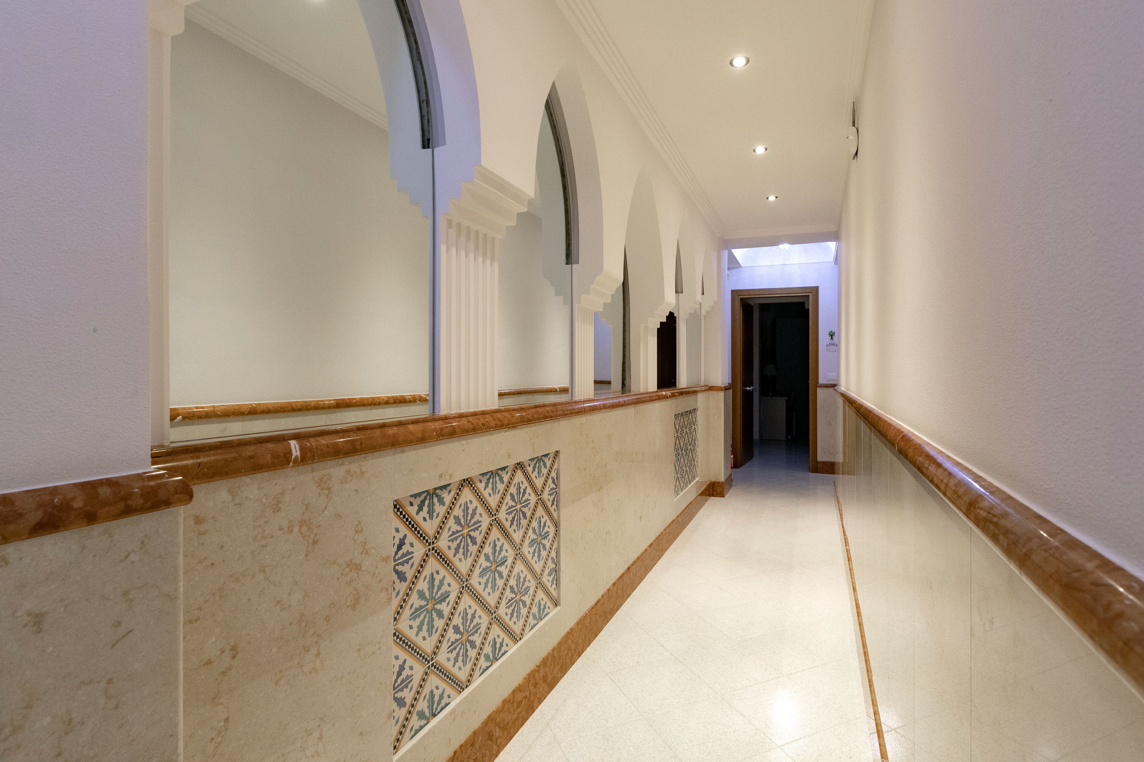 A long, narrow hallway with white walls, a marble floor, and a patterned tile design on the lower wall.