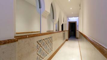 A long, narrow hallway with white walls, a marble floor, and a patterned tile design on the lower wall.