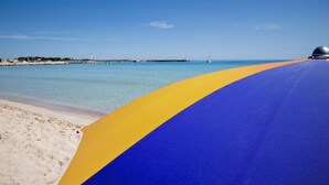 A beach with a clear blue sky, a sandy shore, and a blue and yellow pathway leading to the water. - Hotel Riva Del Sole (San Vito Lo Capo)