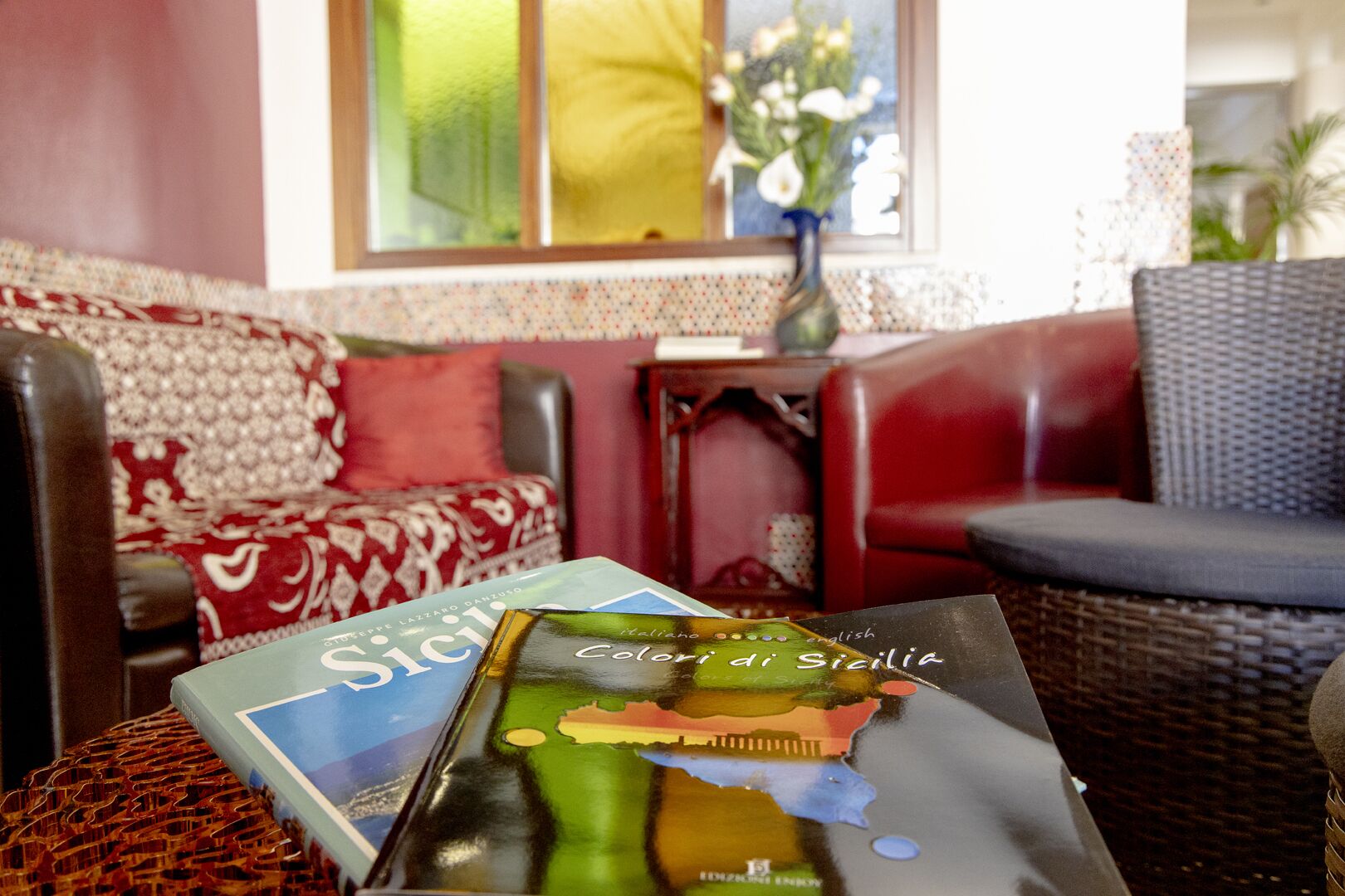 Una habitación con un sofá estampado, una silla de mimbre y una mesa con un libro titulado 'Colori di Sicilia'.