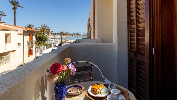 A balcony with a table set for breakfast, overlooking a beach and buildings. - Hotel Riva Del Sole (San Vito Lo Capo)