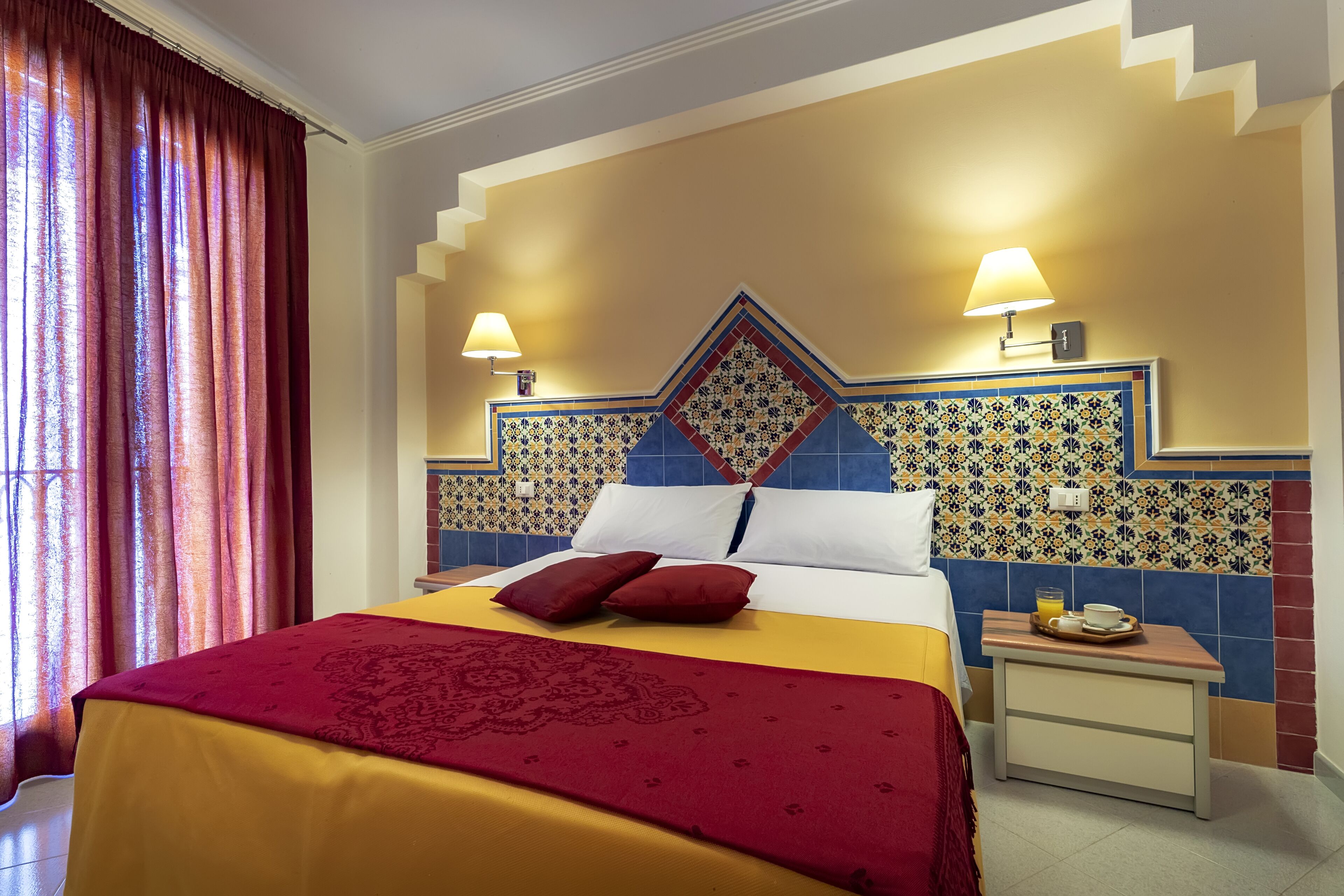 A hotel room with a bed, two bedside lamps, a nightstand with a tray of breakfast items, and a decorative wall tile pattern.