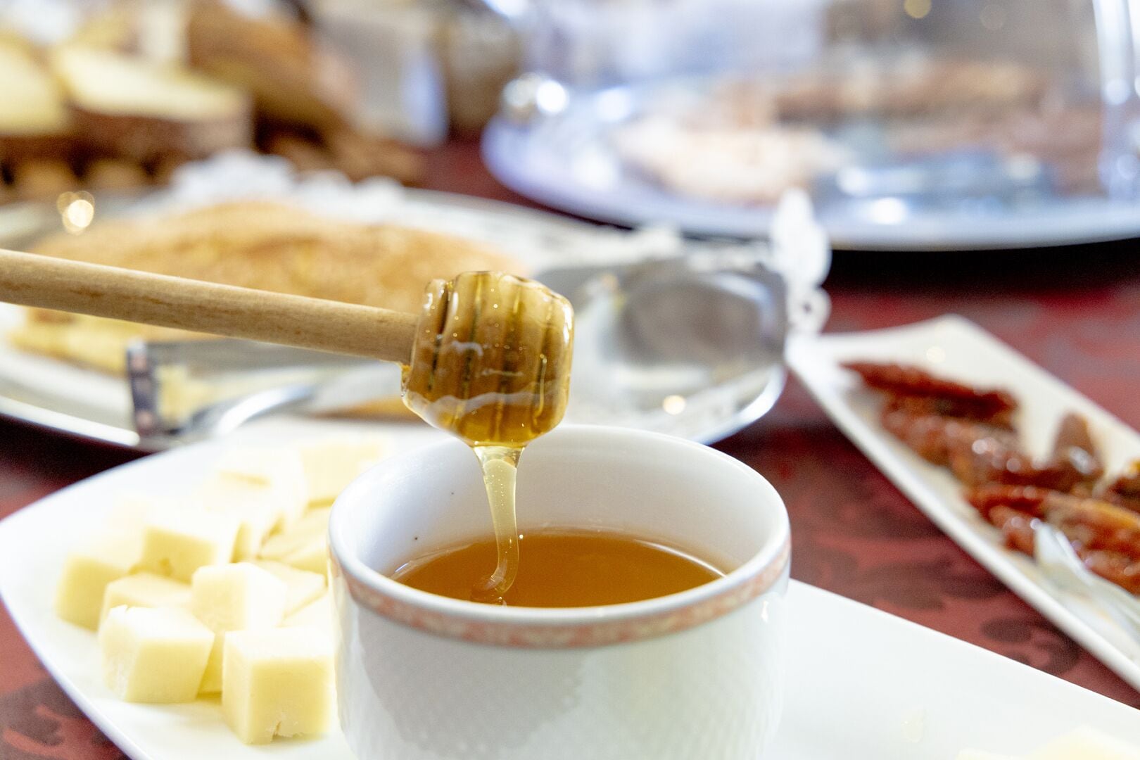 A table with a white cup of tea, honey being drizzled, and plates of food including cheese and bacon.