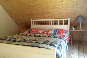 Un dormitorio con techo de madera, una cama blanca con colcha, una mesita de noche con lámpara y una pequeña mesa con un jarrón.