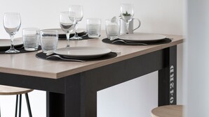 Un comedor con un reloj de pared, una mesa dispuesta con platos, vasos y cubiertos, y una silla.