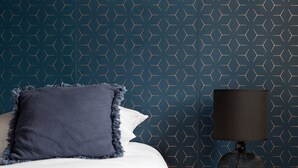 Una cama bien hecha con una manta azul oscuro y almohadas, una lámpara negra sobre una mesita de noche de madera y una pared con un diseño geométrico.