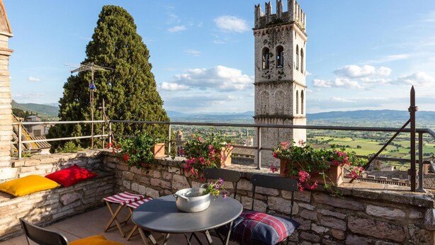 A stone balcony with a table, chairs, and potted plants. A tall tower with arched windows is visible in the background. - Room En Suite Corte Assisi (Assisi)