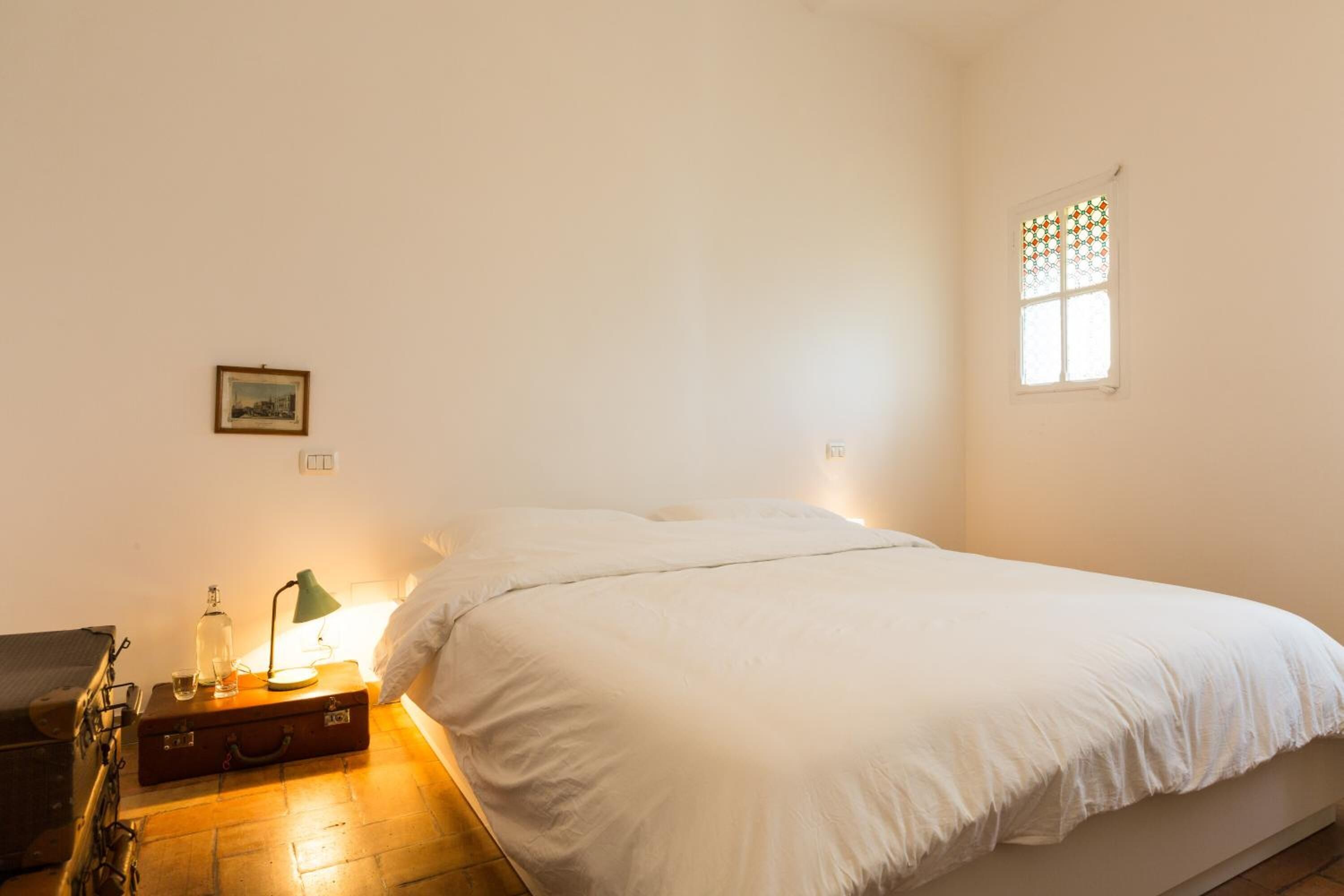Quarto com cama, mala, abajur e uma mesinha com garrafa e copos.