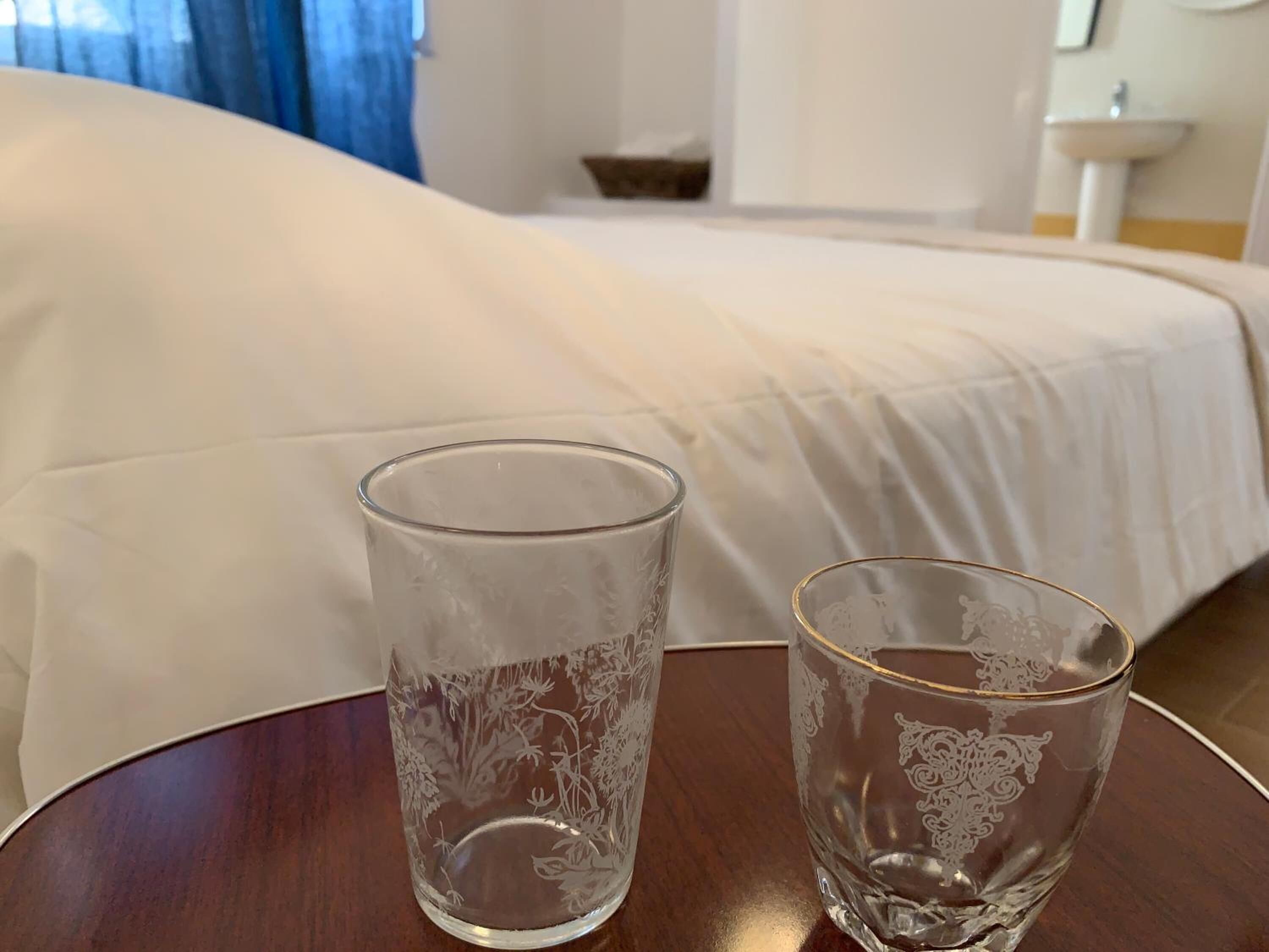 Zwei klare Glasbecher auf einem Holztisch in einem Hotelzimmer, im Hintergrund ein Bett.