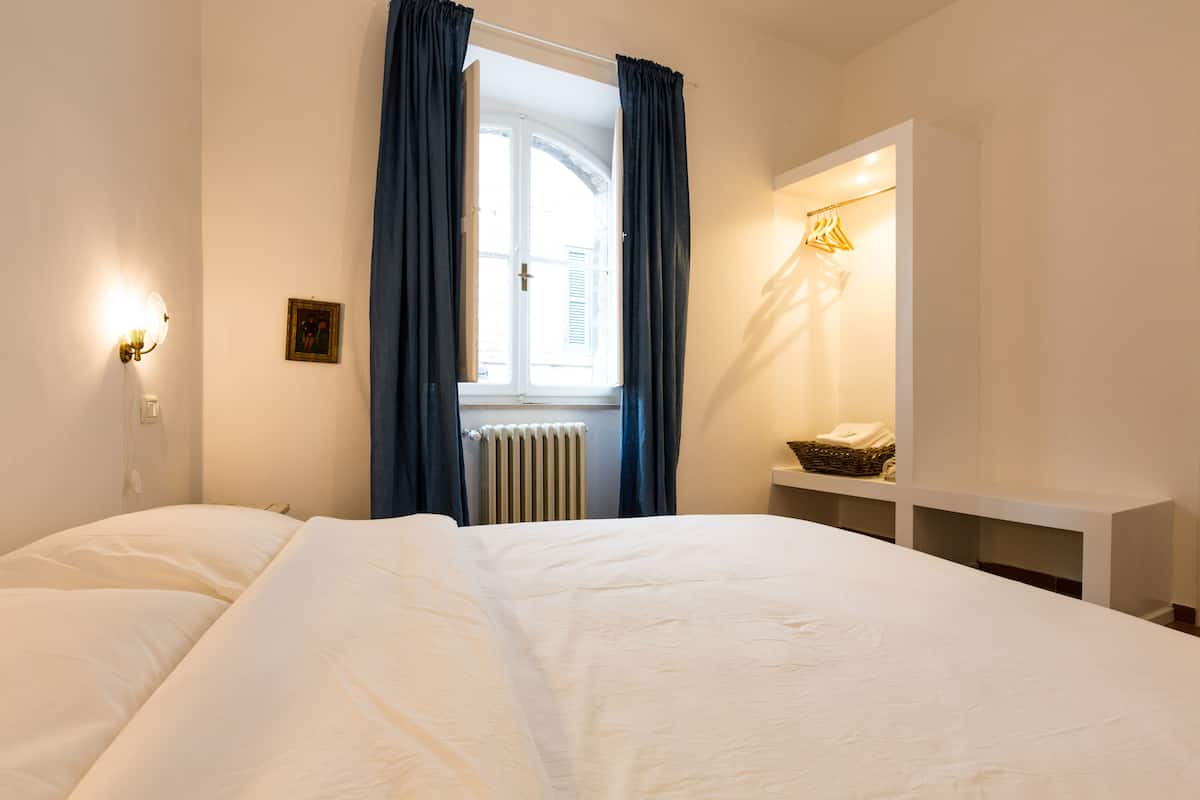 A hotel room with a large bed, a bedside table with a lamp, a window with curtains, and a radiator.