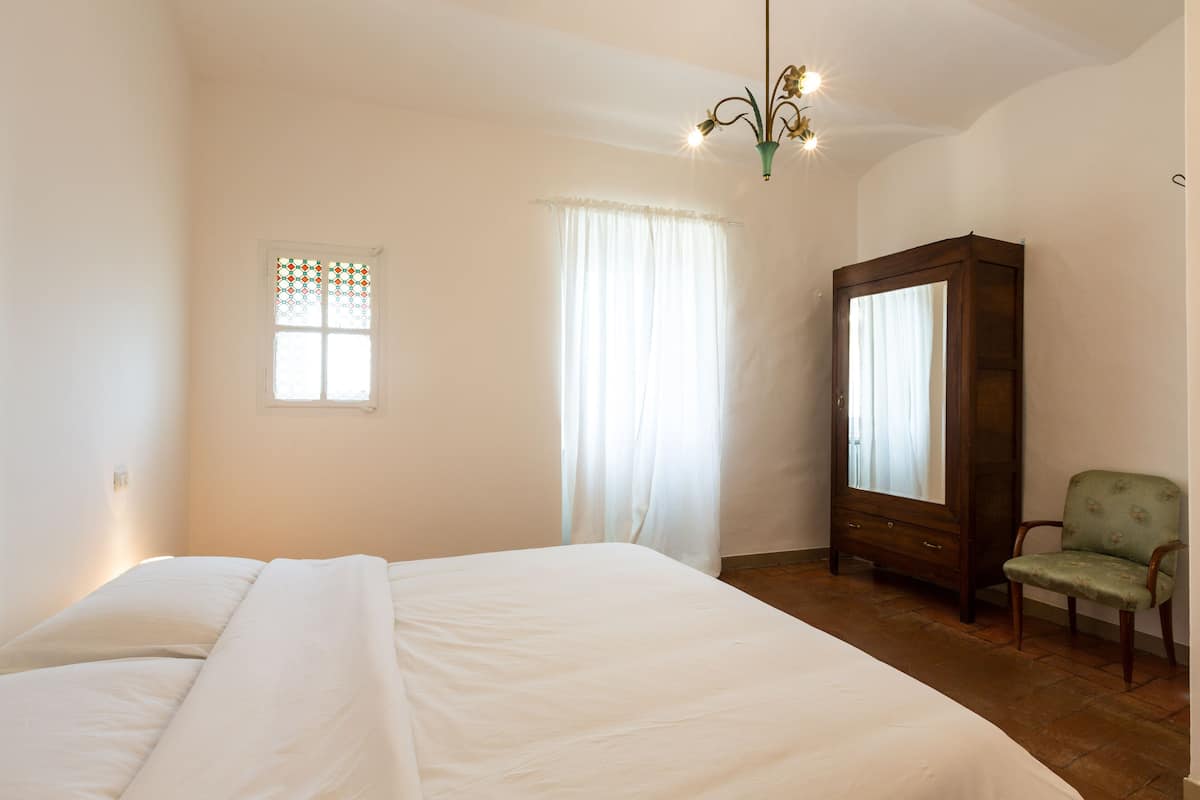 A bedroom with a large bed, a wooden wardrobe, a chair, and a window with curtains.