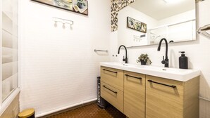 A bathroom with a white tiled wall, a wooden vanity with a black faucet, a mirror, and a small plant on the counter. - Les Embruns - Appt 2 ch avec parking (Saint-Cyprien)