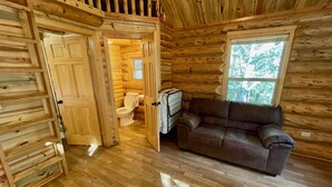 A log cabin interior with a wooden floor, a sofa, a window, and a door leading to a bathroom. - Secluded Creekside Cabin (Coaldale)