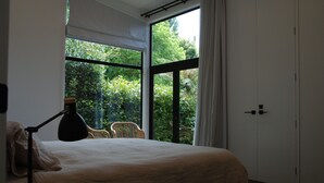 A bedroom with a large window, a bed, a chair, and a desk with a lamp.