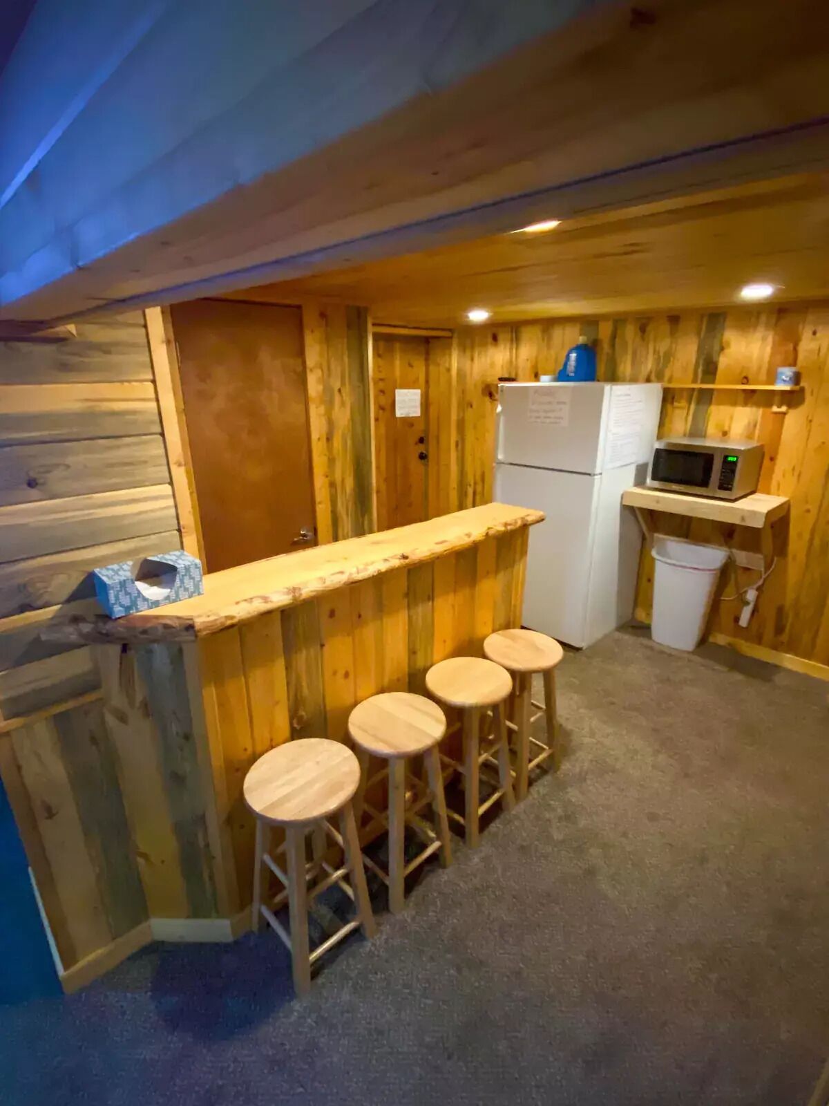 A compact kitchen with wooden walls, a white refrigerator, a microwave on a shelf, and three stools.