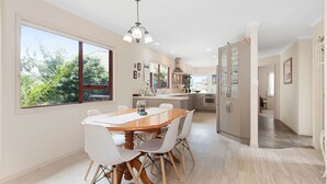 A modern kitchen with a central island, large windows, and a dining area with a wooden table and white chairs. - Sandy Feet - Papamoa Beach Holiday Home (Papamoa)