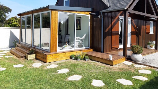 Una casa moderna con un patio con paredes de vidrio, terraza de madera y un pequeño jardín.