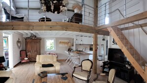 Un amplio salón con techo alto, vigas de madera, un sofá, un sillón, un televisor y una cocina al fondo.