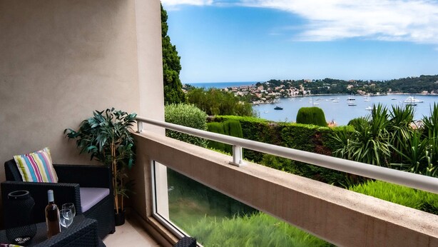 Un balcon donnant sur une ville côtière, un canapé en osier et une table avec une bouteille et un verre.