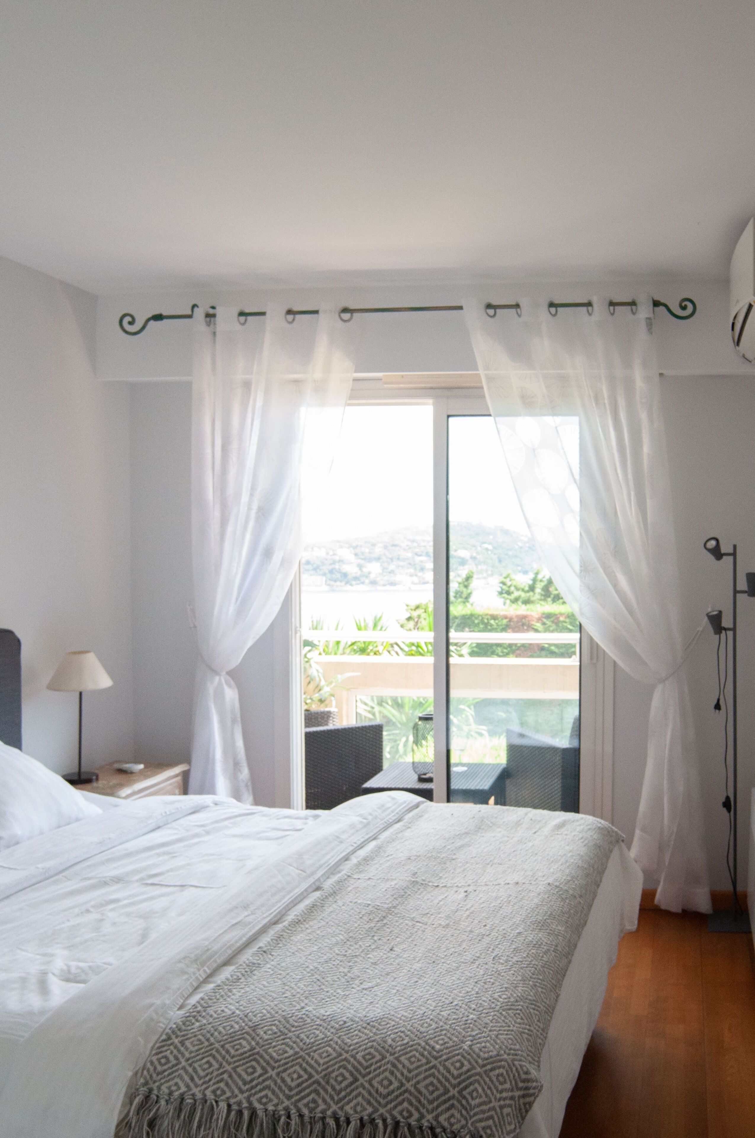 Ein Schlafzimmer mit einem Bett, einem Nachttisch, einer Lampe und Blick auf einen Balkon mit Gartenmöbeln.