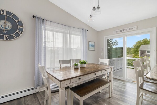 A dining area with a wooden table, white chairs, a bench, a clock, and a view of the outdoors.