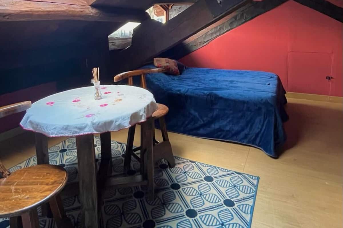 Ein kleines Dachzimmer mit schrägem Giebel, einem Einzelbett, einem kleinen Tisch mit einer Vase und einem gemusterten Teppich.