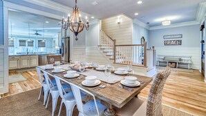 A spacious dining area with a large wooden table set for a meal, surrounded by white chairs. A chandelier hangs above, and a staircase leads to an upper level. - Exclusive Pet-Friendly Watersound West Beach Retreat with Electric Cart, Bikes, Pool & Beach Access (Santa Rosa Beach)
