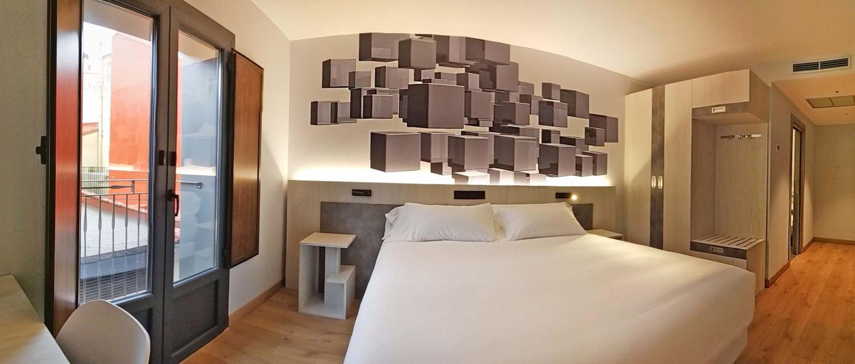 Una habitación de hotel moderna con una cama grande, una mesita de noche pequeña, una lámpara de pared y una puerta corrediza que da a un balcón.