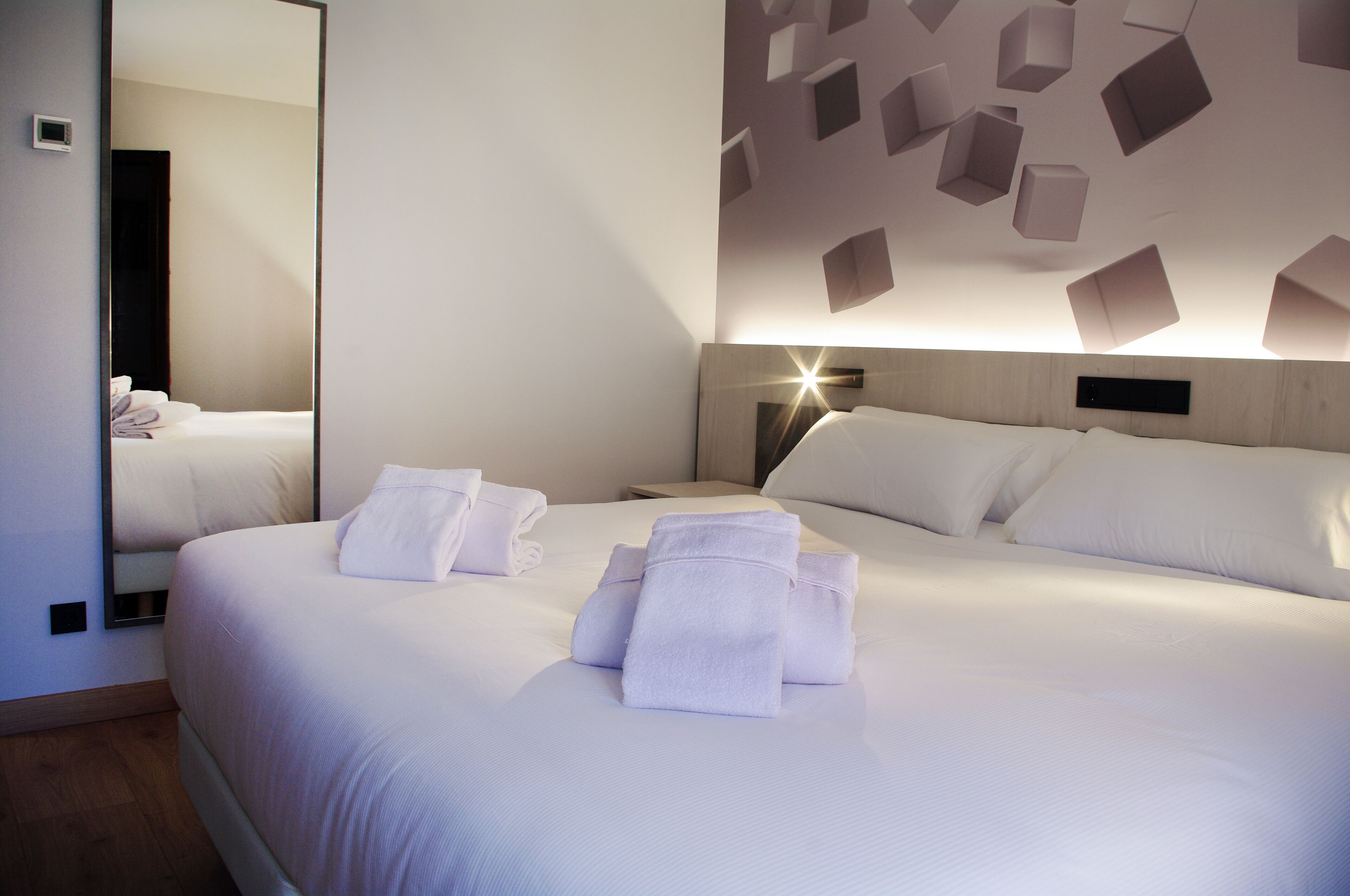 A hotel room with a large bed, a bedside table, a mirror, and a wall with geometric light fixtures.