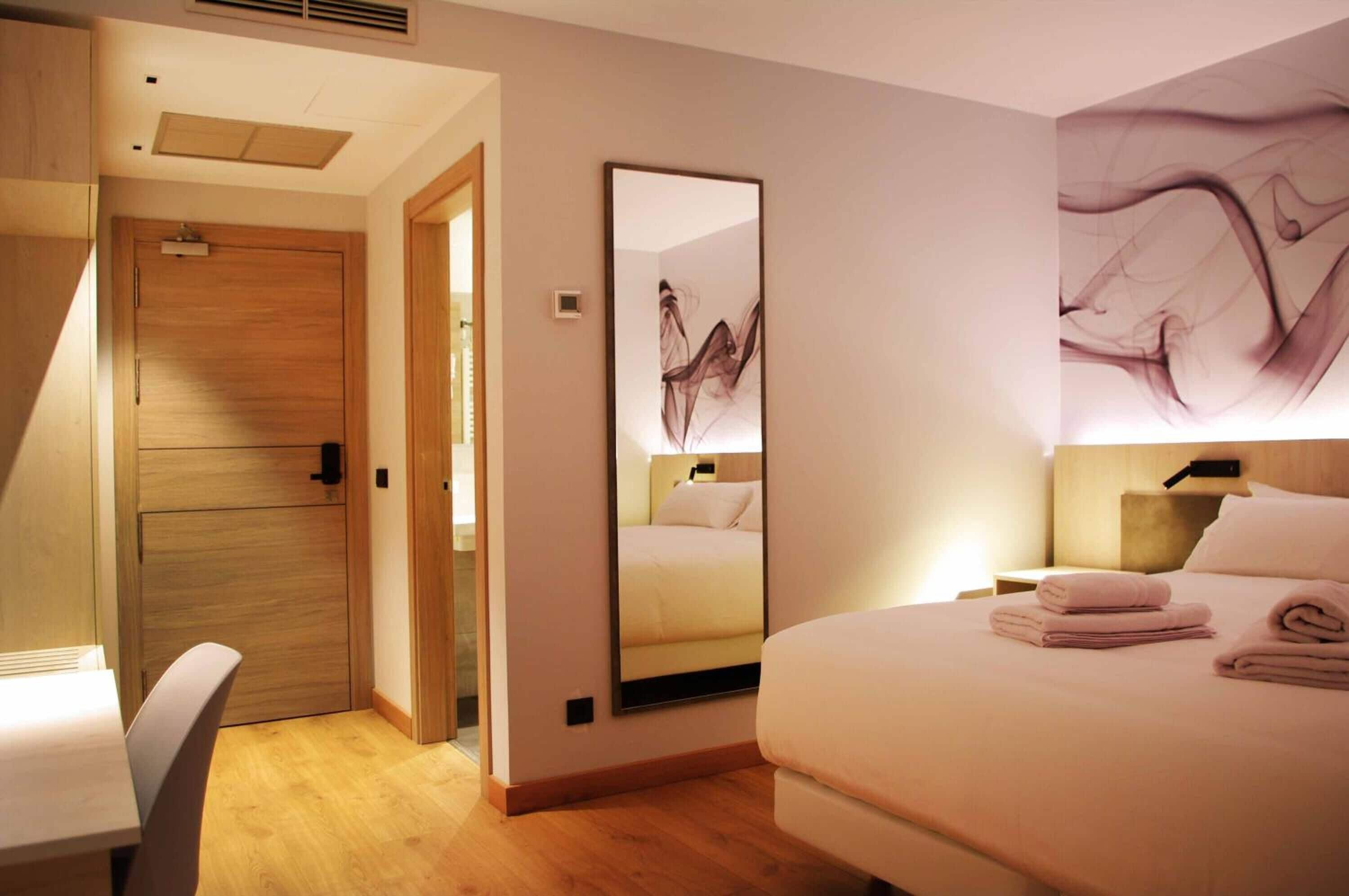 Hotelkamer met een bed, nachtkastje, stoel, houten garderobe, spiegel en een muur met abstracte kunst.