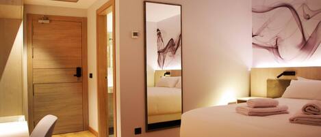 Hotelkamer met een bed, nachtkastje, stoel, houten garderobe, spiegel en een muur met abstracte kunst.