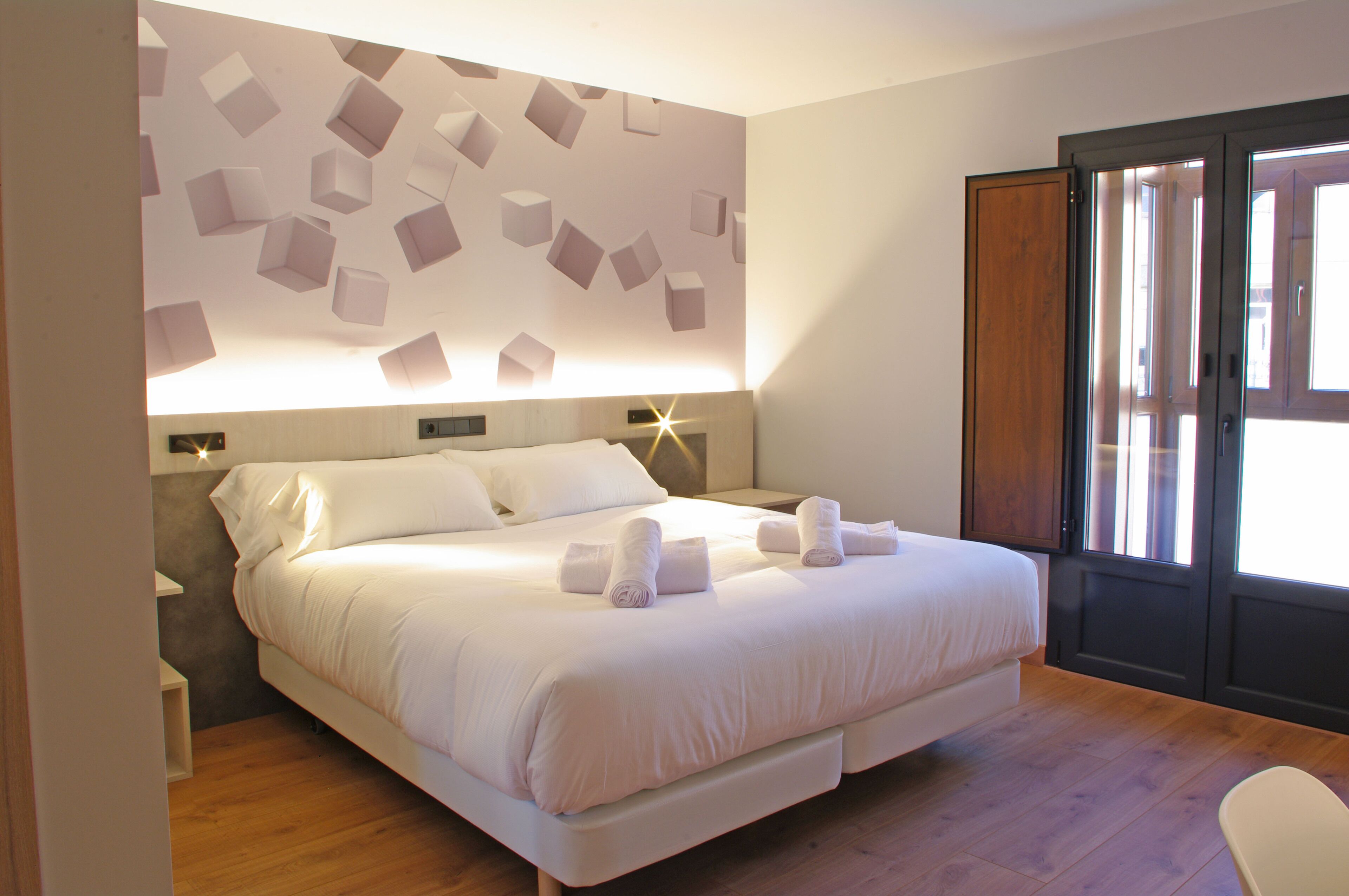 A bedroom with a large bed, a headboard with a geometric light fixture, a wooden door, and a view of the outdoors.
